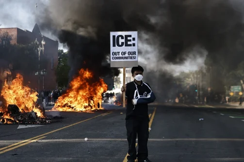 a protester holds a sign reading ice out of our communities as burning waymo cars line the street